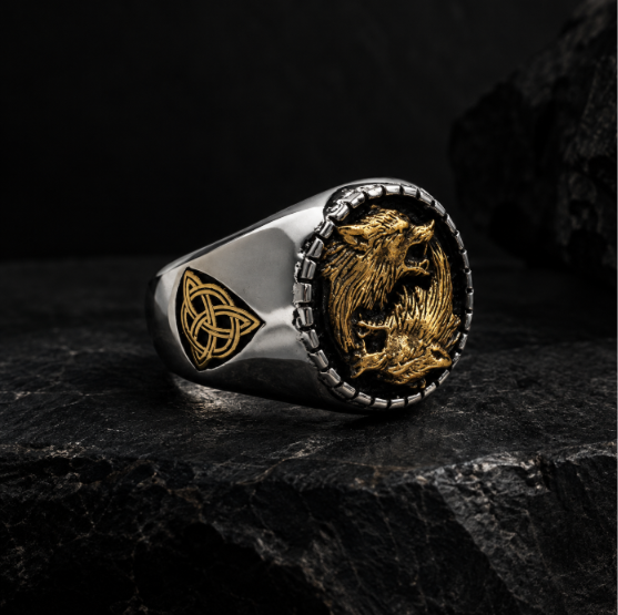 A silver-toned signet ring with a gold-toned double-headed eagle emblem on the face and a gold Celtic knot triangle on the band, displayed on a dark textured stone surface.