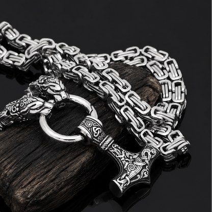 A silver-toned Viking-style chain necklace with a hammer pendant and lion head ring, displayed on a dark wood block against a black reflective background.