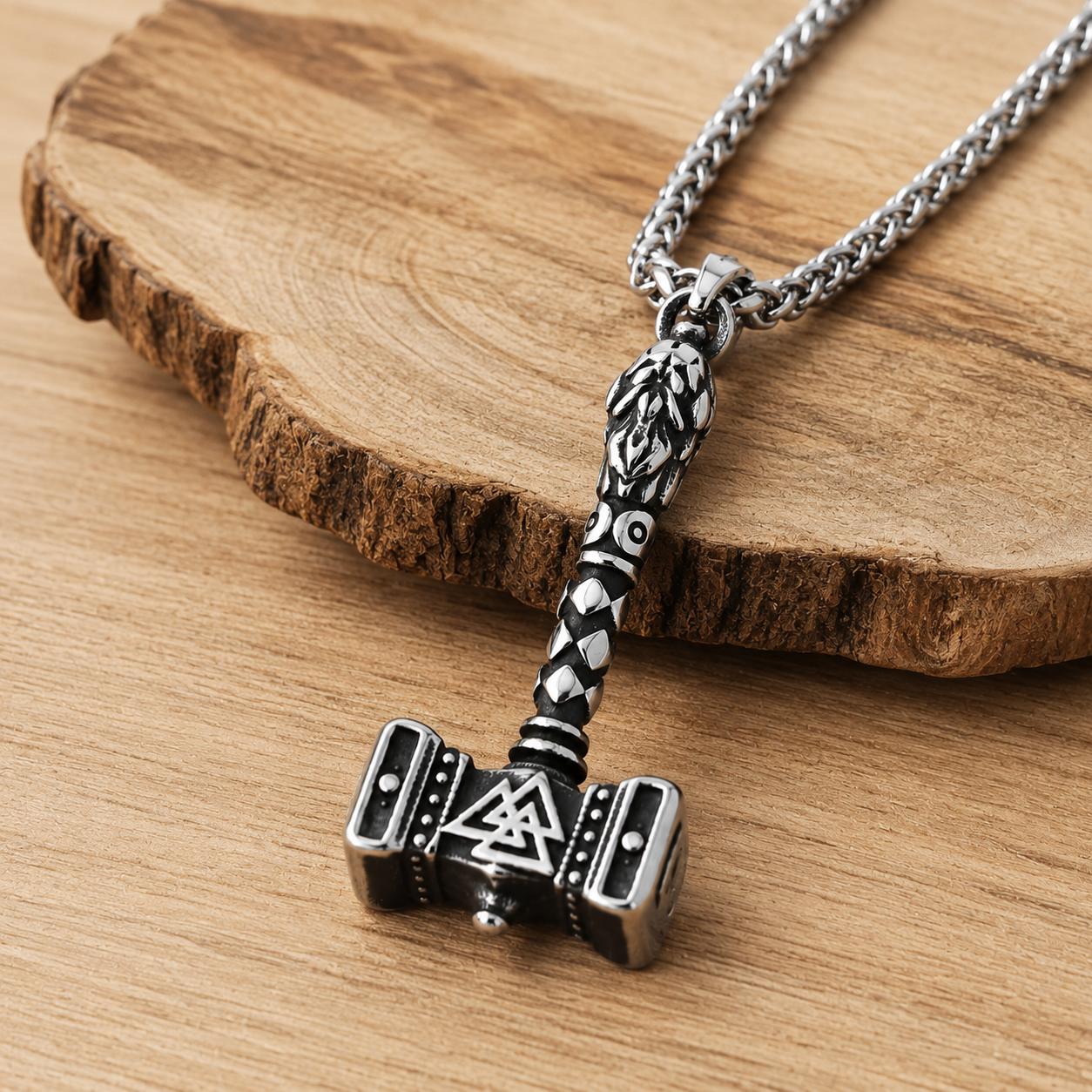 A silver-toned metal pendant necklace shaped like a hammer, resting on a rustic wood slice over a light wood surface. The pendant has engraved black details and a chain is partially visible at the top left.